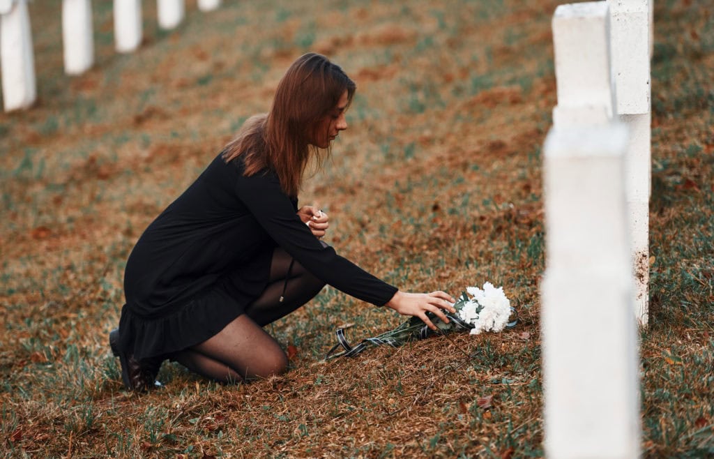 Gives respect by putting flowers. Young woman in black clothes visiting cemetery with many white crosses. Conception of funeral and death