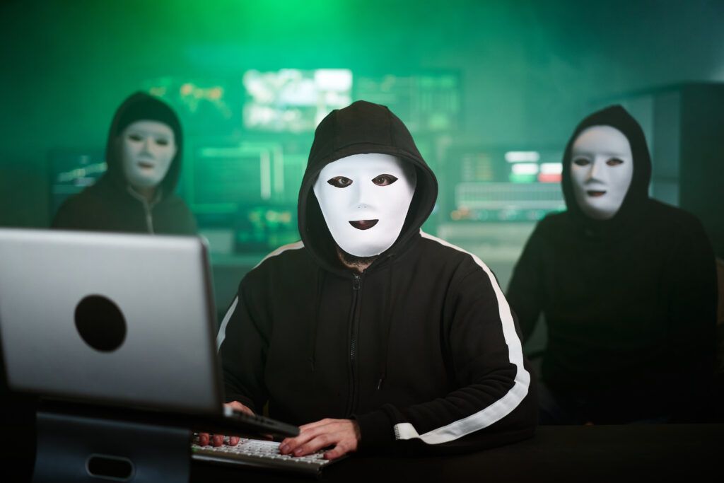 Three masked individuals in hooded sweatshirts working on computers in a dark, tech-lit room, representing cybercrime and privacy violations online