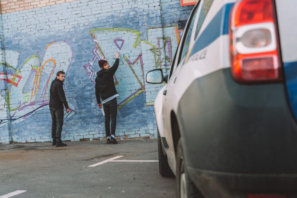 Two individuals spray-painting graffiti on a wall while a police vehicle approaches, highlighting legal consequences of vandalism