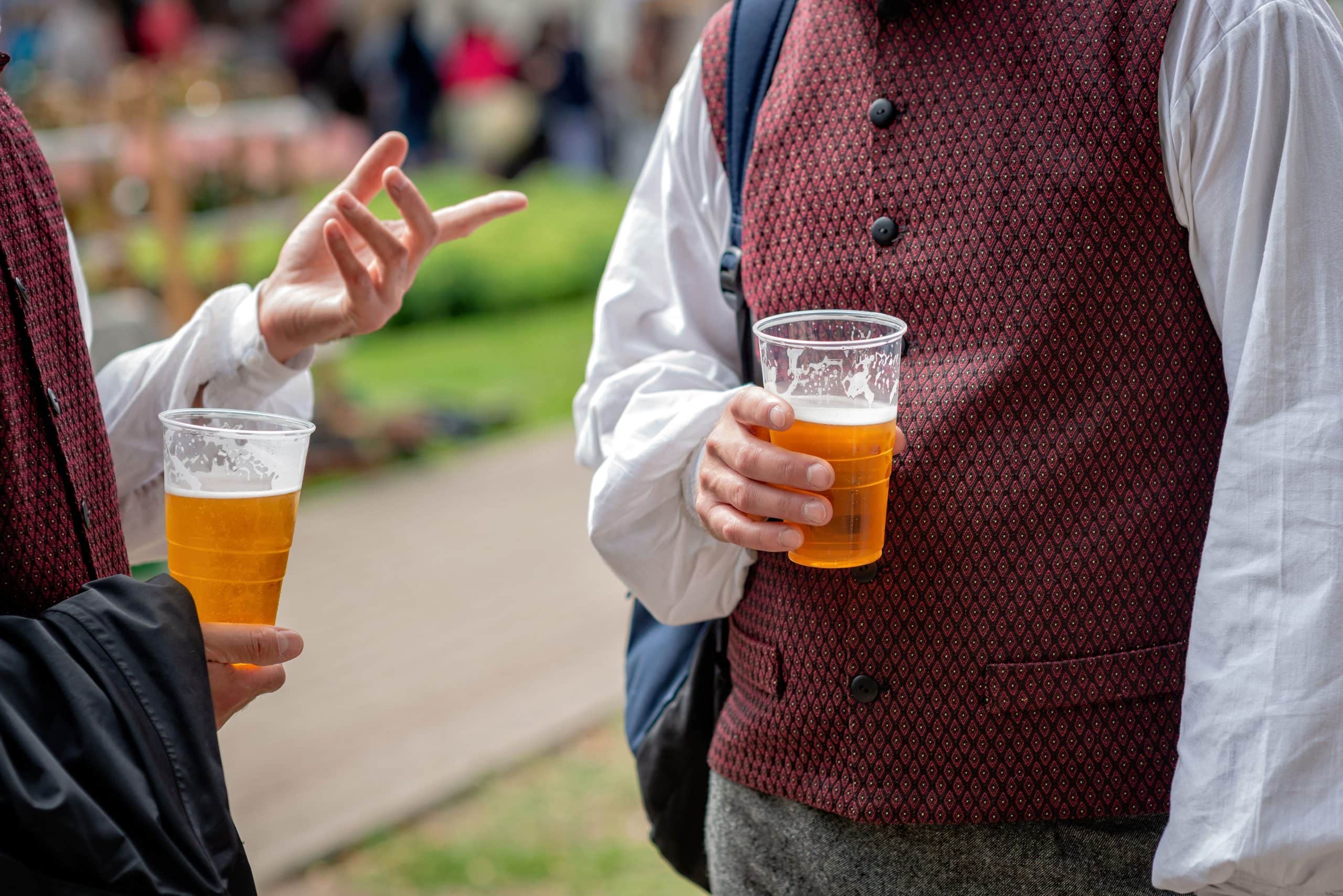 Two men in folk costumes talk and drink beer from plastic glasse