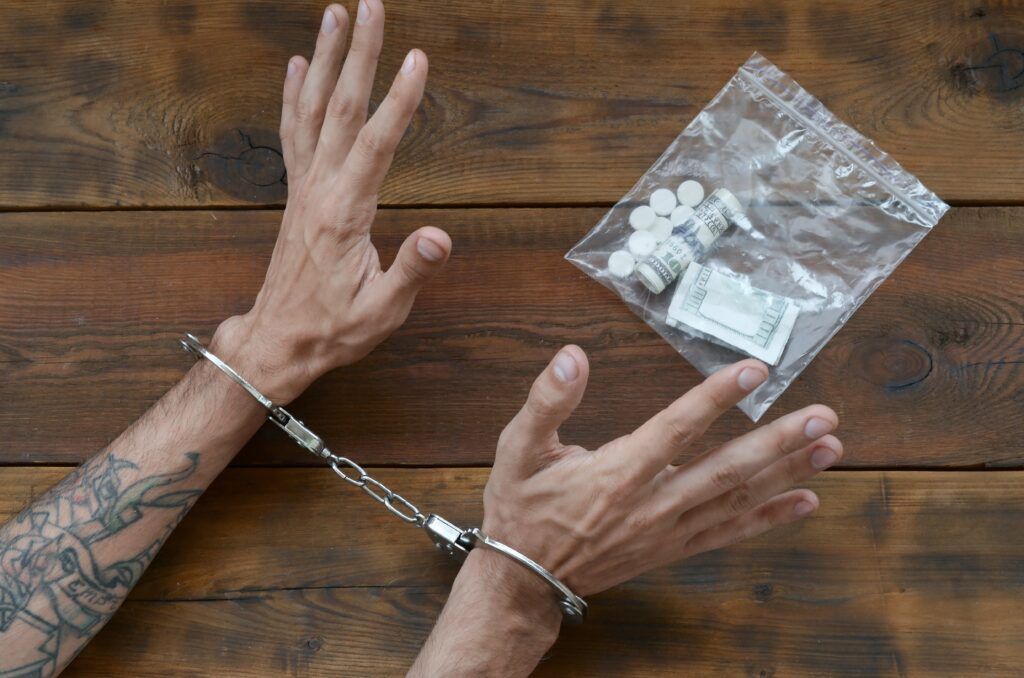 Handcuffed hands on a wooden table beside a clear bag of pills and cash, representing possession for the purpose of trafficking charges and penalties in Canada.
