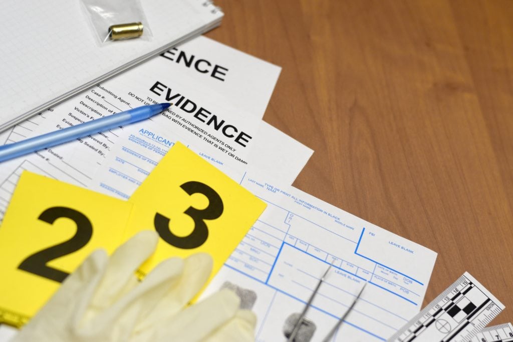 Close-up of forensic evidence documents, numbered crime scene markers, a bullet casing, and fingerprint forms on a wooden table.